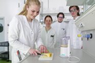 Personen in weißer Kleidung stehen um einen Tisch, eine Person schneidet Käsestück