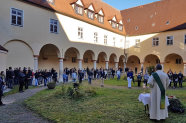 Mann in Priestergewand steht vor Personen im Innenhof eines Gebäudes